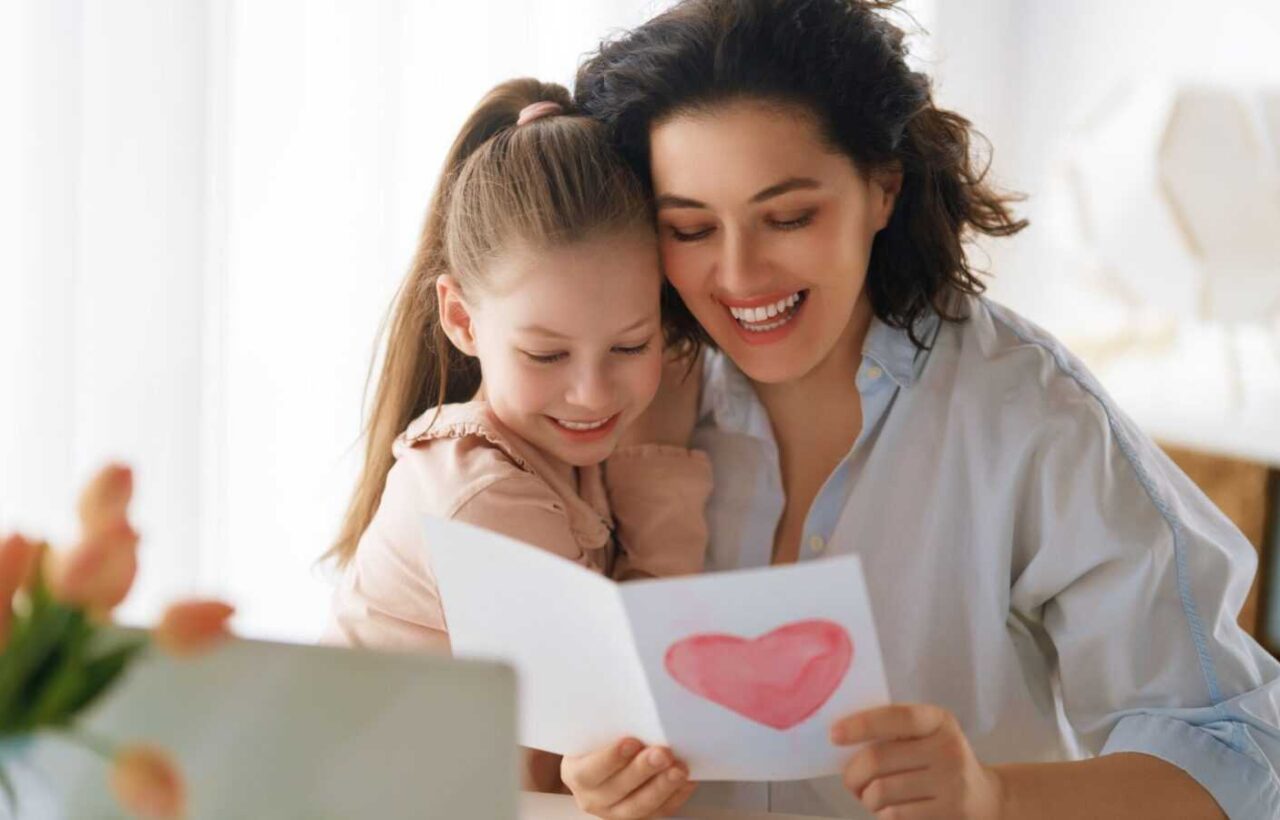 Mother and young daughter smiling while reading a handmade heart card at home, representing family love and support for working moms in Canada.