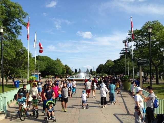 Families and visitors stroll along a sunny Toronto park promenade with Canadian flags, strollers, trees, and a fountain in the distance.
