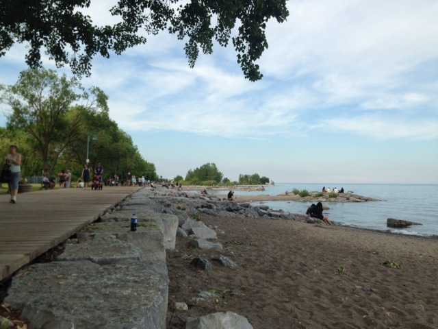 Toronto waterfront beach with a boardwalk, rocky shoreline, people relaxing by Lake Ontario, trees, and blue summer sky.