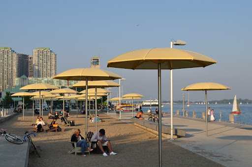 People relax under yellow umbrellas along Toronto's waterfront, with sandy paths, lake views, sailboats, and city buildings in summer.