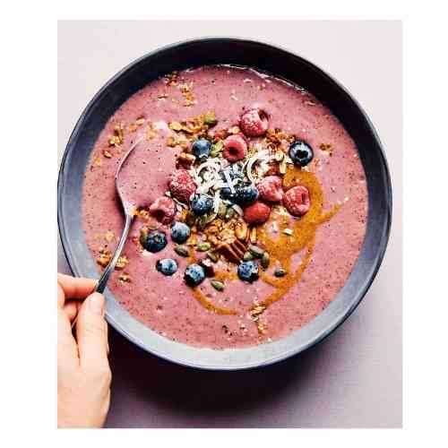 Overhead view of a pink berry smoothie bowl topped with blueberries, raspberries, granola, coconut, seeds, and a nut butter drizzle.