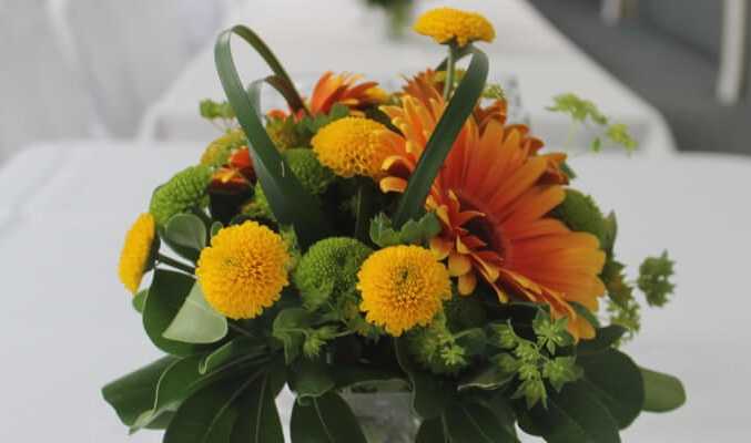 Bright Mother's Day floral arrangement with orange gerbera daisies, yellow billy buttons, green mums, and glossy leaves on a white table.