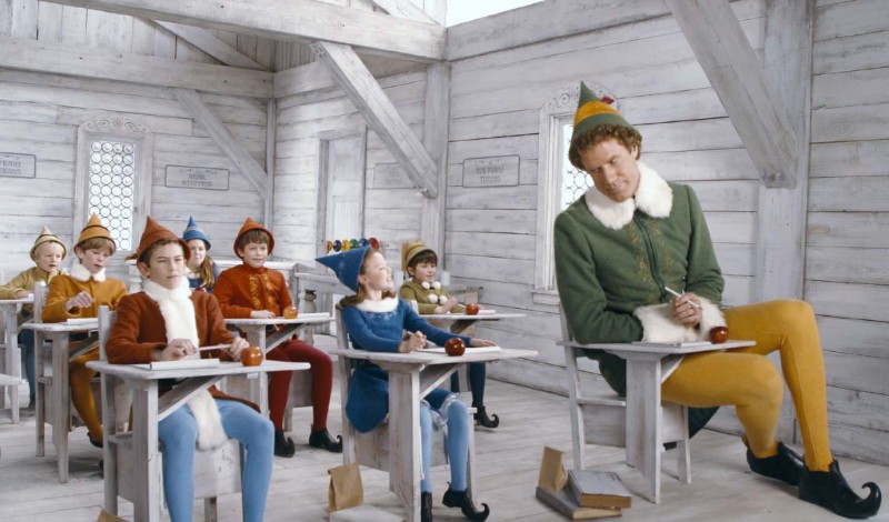 Buddy the Elf sits in a tiny classroom desk with elf children in a snowy North Pole classroom from the holiday movie Elf.