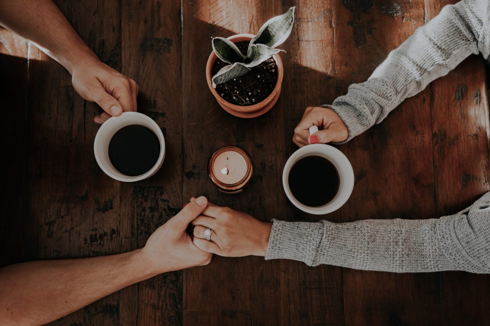 Overhead view of a couple holding hands beside coffee mugs, a candle, and a small potted plant on a wooden table at home.