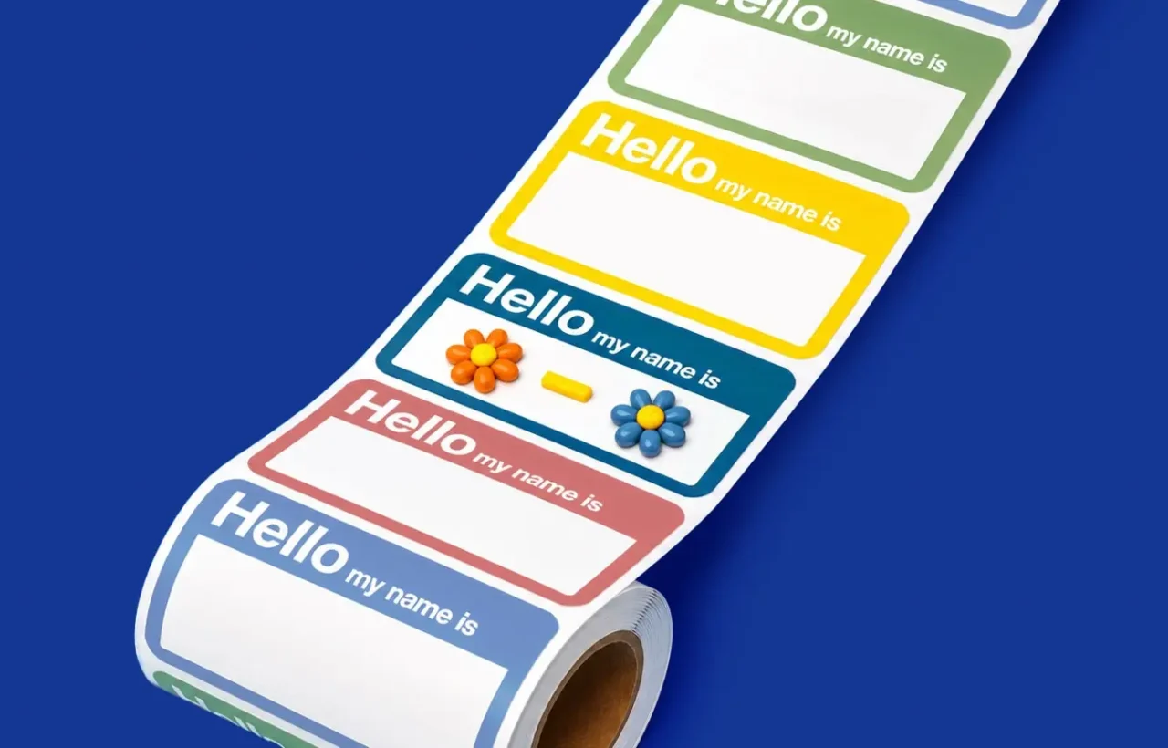 Roll of colorful “Hello my name is” name tag stickers with a hyphen and flower decorations on a bright blue background.