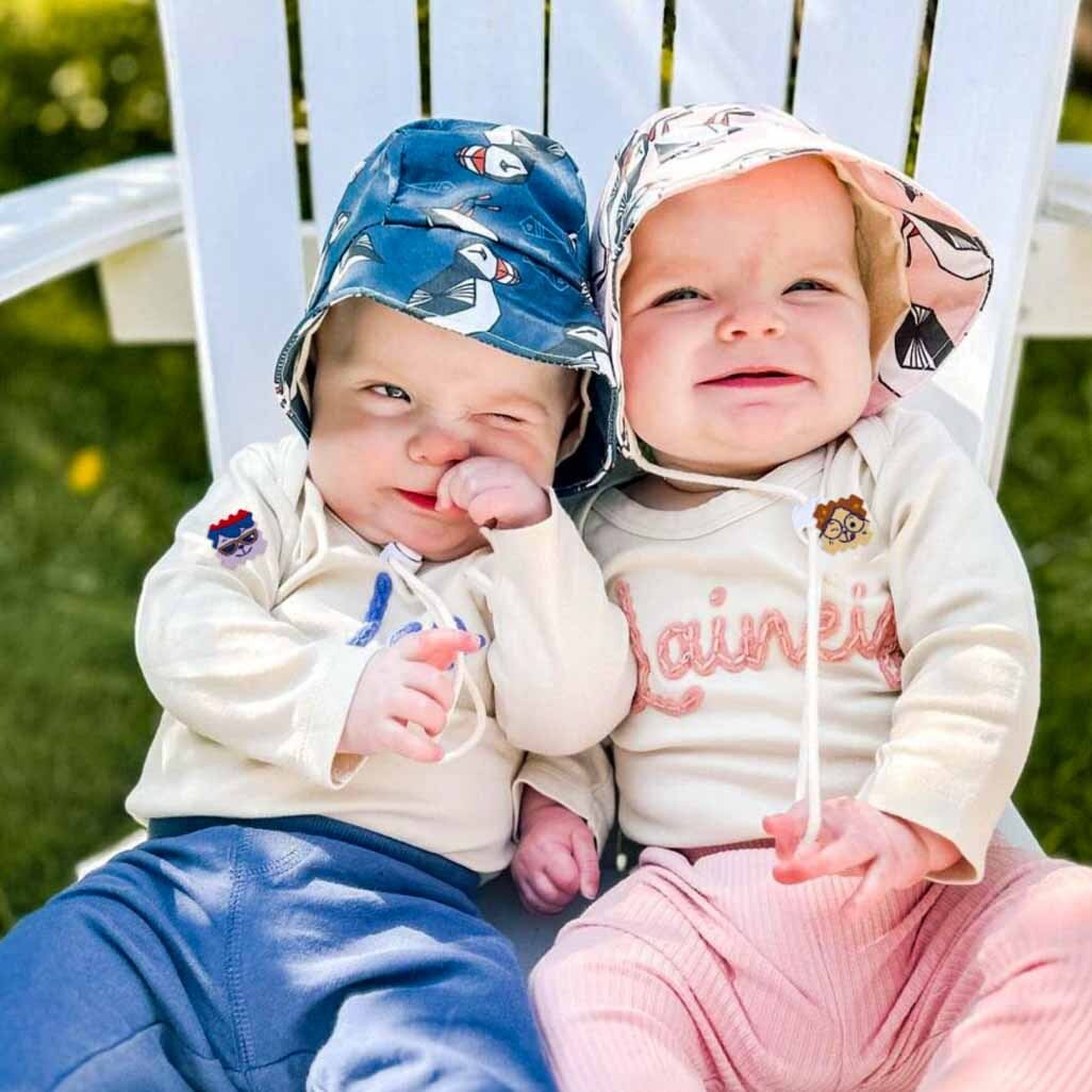 Two babies in patterned sun hats sit together on a white outdoor chair, wearing light outfits with colorful patches on a sunny day.