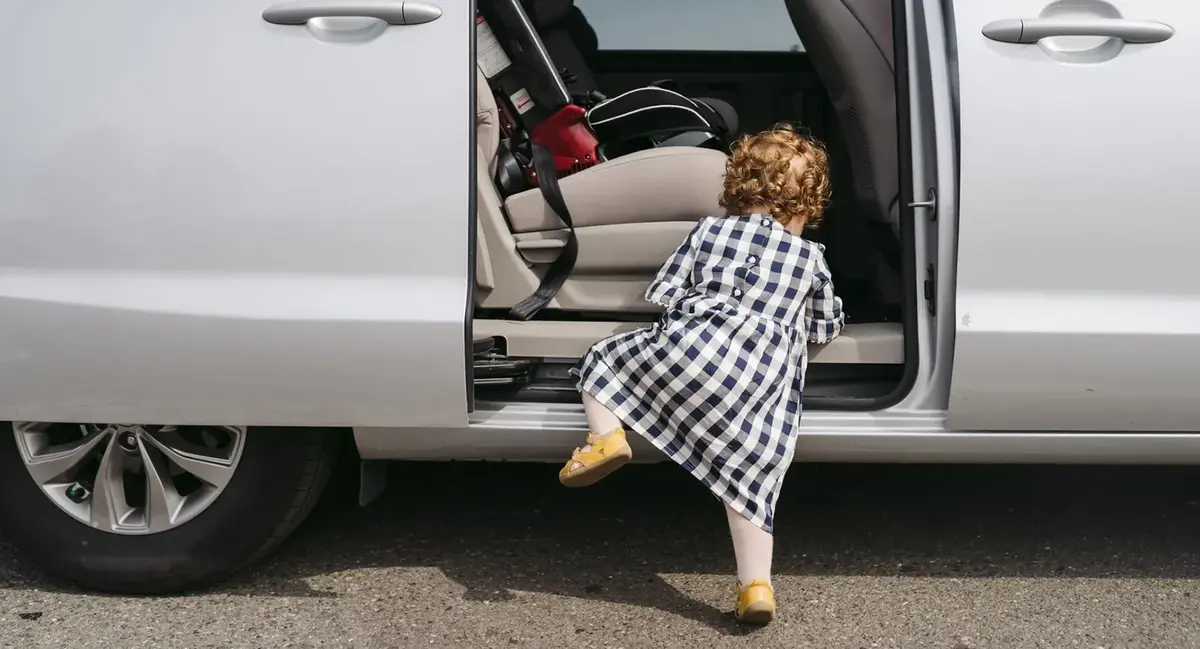 Toddler in a checkered dress climbing into a silver family SUV with an open door, showing child access and vehicle space considerations.