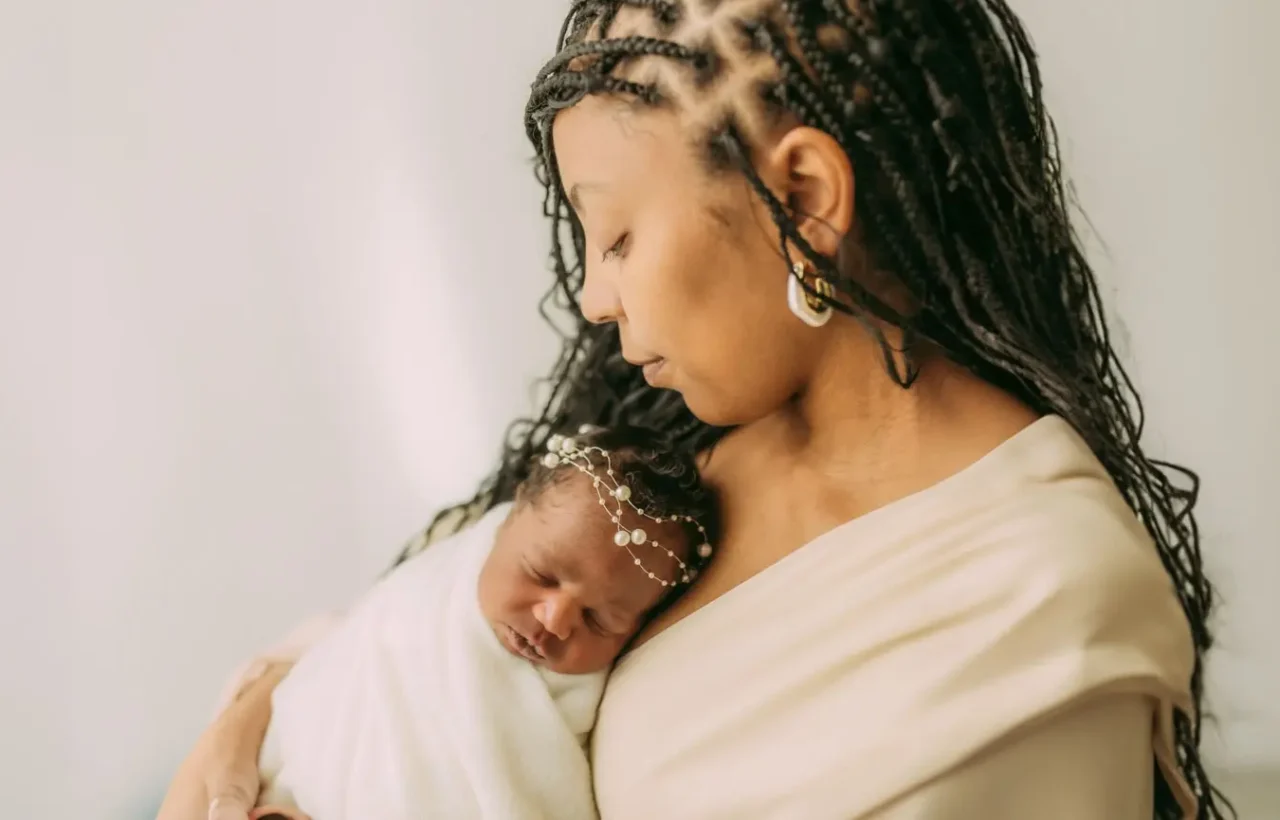 Mother with braided hair gently cradles her sleeping newborn wrapped in a white blanket, a tender portrait for newborn health and hearing care.