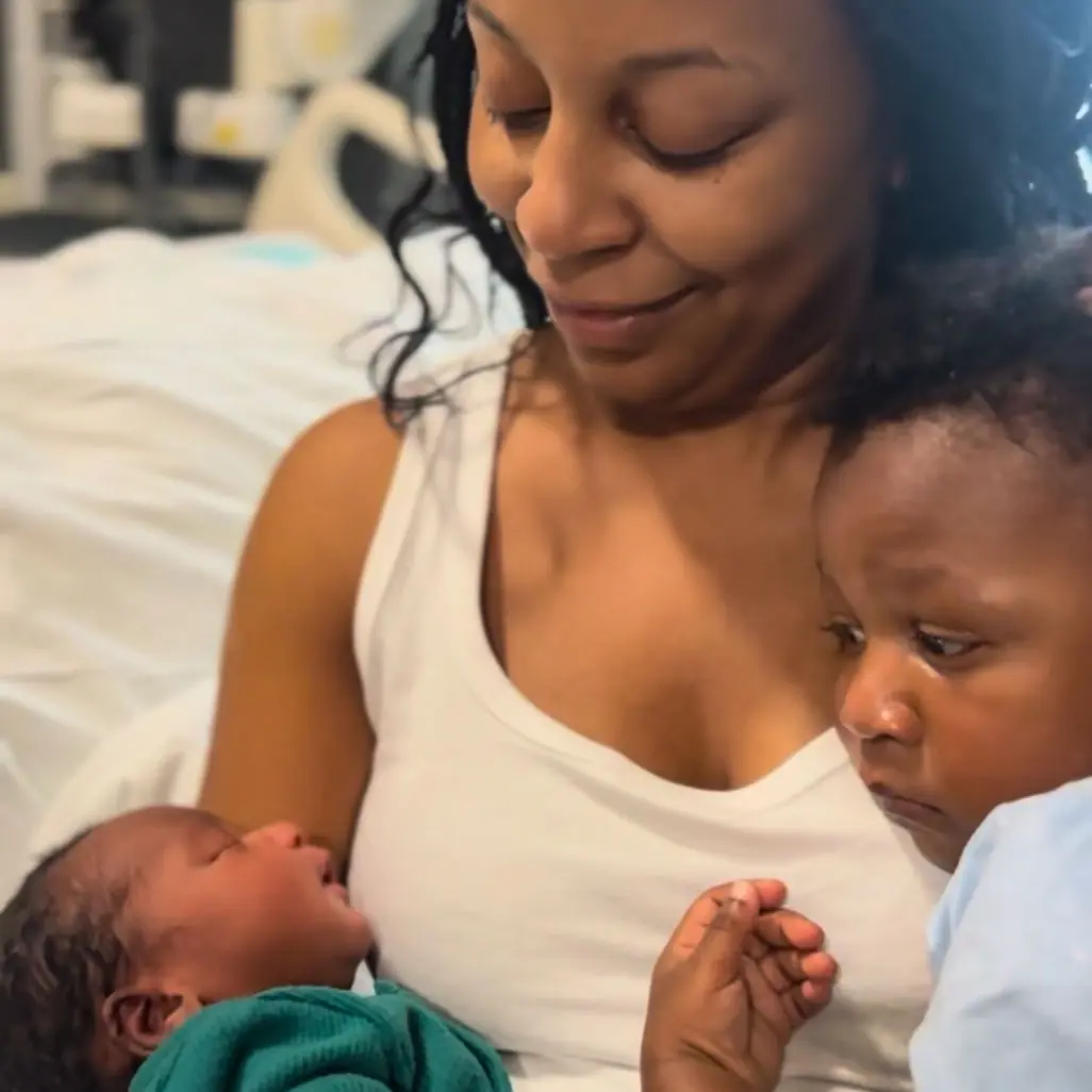 Mother in a hospital bed cradles a newborn while an older child looks on, capturing a quiet family moment after birth.