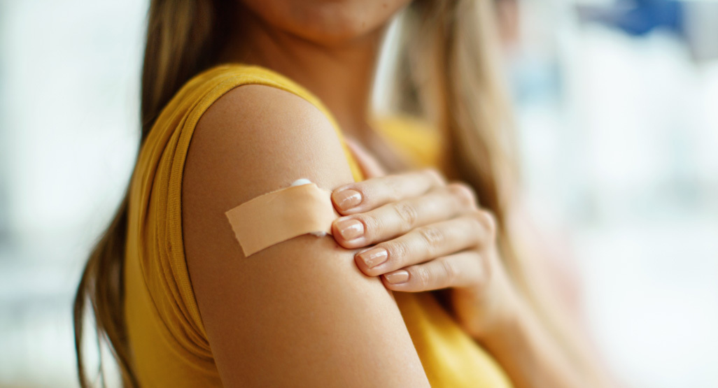 Pregnant woman in a yellow top touches a bandage on her upper arm after receiving an RSV vaccine shot.