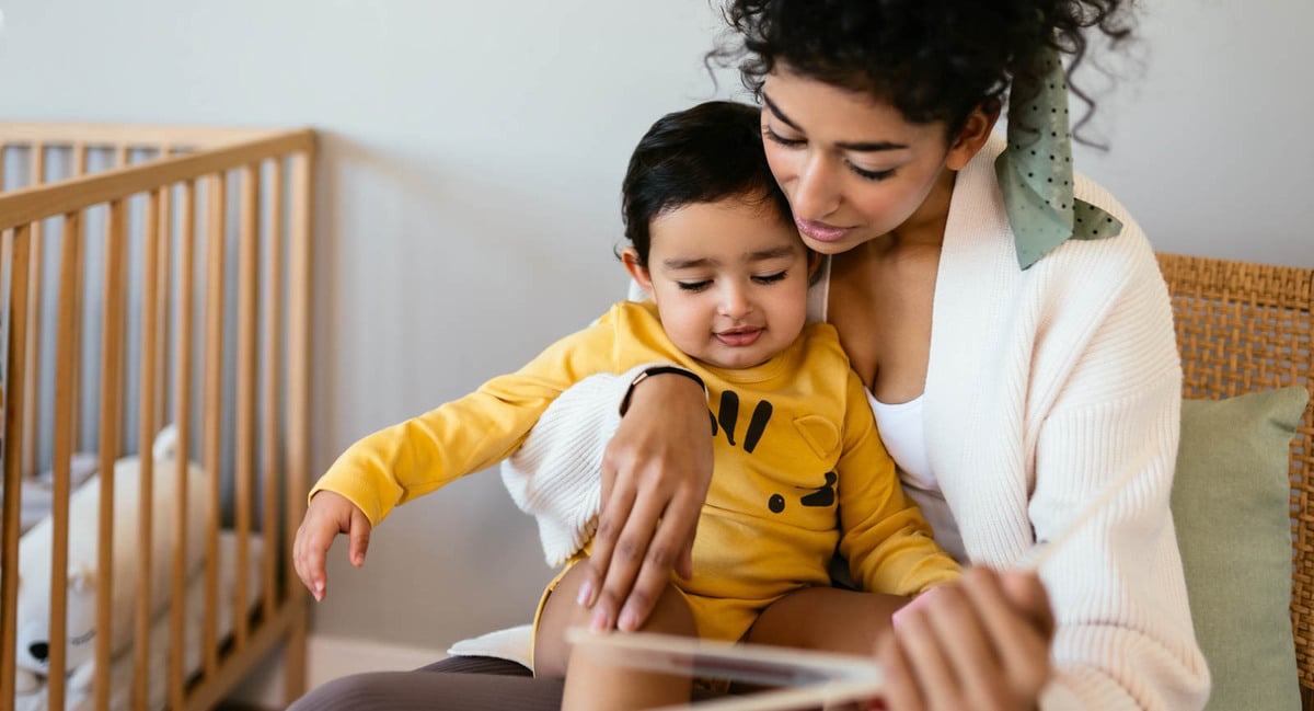 Mother cuddles her baby while reading a book together in a nursery, supporting early language development and baby hearing awareness.