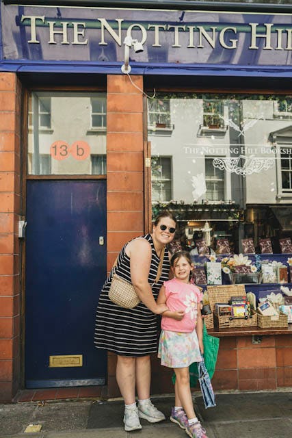 A woman and young girl smiling outside The Notting Hill Bookshop in London, standing by a blue door and shop window on a sunny family trip.