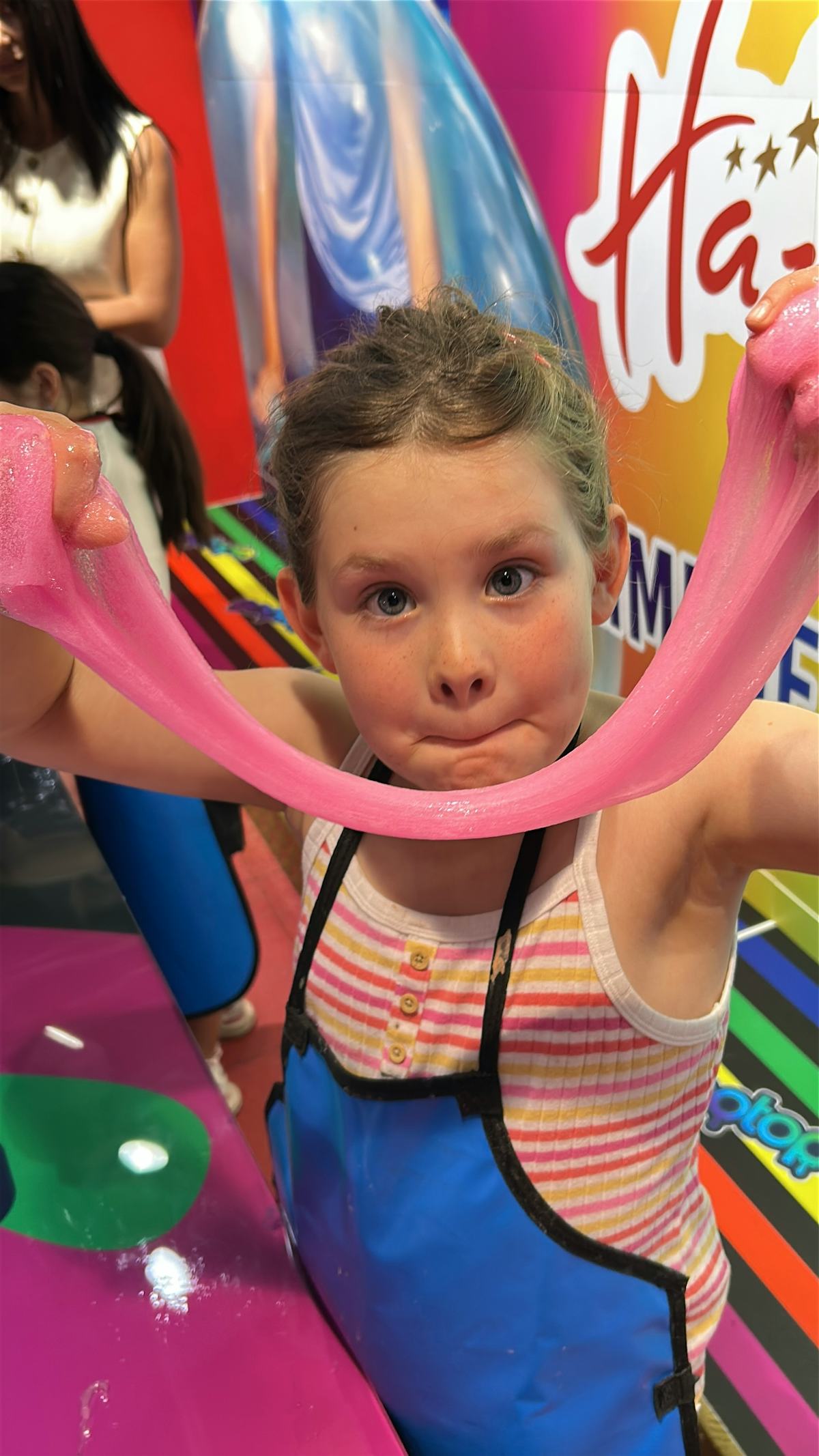 Child stretches bright pink slime at Hamleys toy store during a family trip to London, wearing a blue apron in a colorful play area.