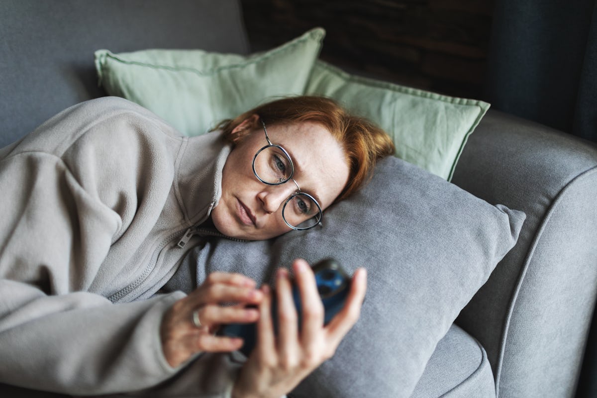 Woman with glasses lying on a couch and looking at her phone, conveying exhaustion, stress, and the need for supportive texts from friends