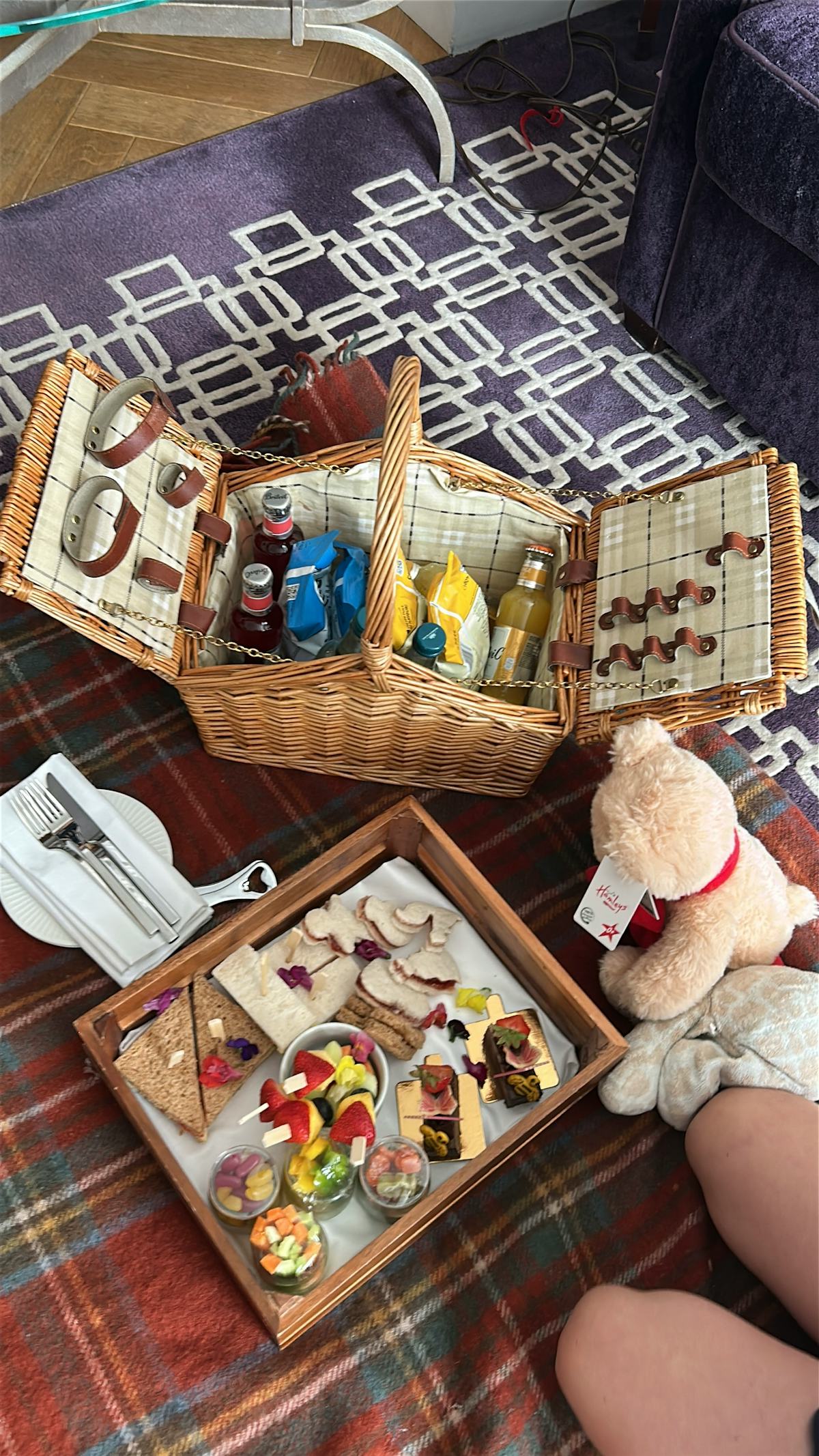 Hotel room picnic setup with wicker basket, teddy bear, tray of snacks, drinks, plates and blanket during a family London trip.