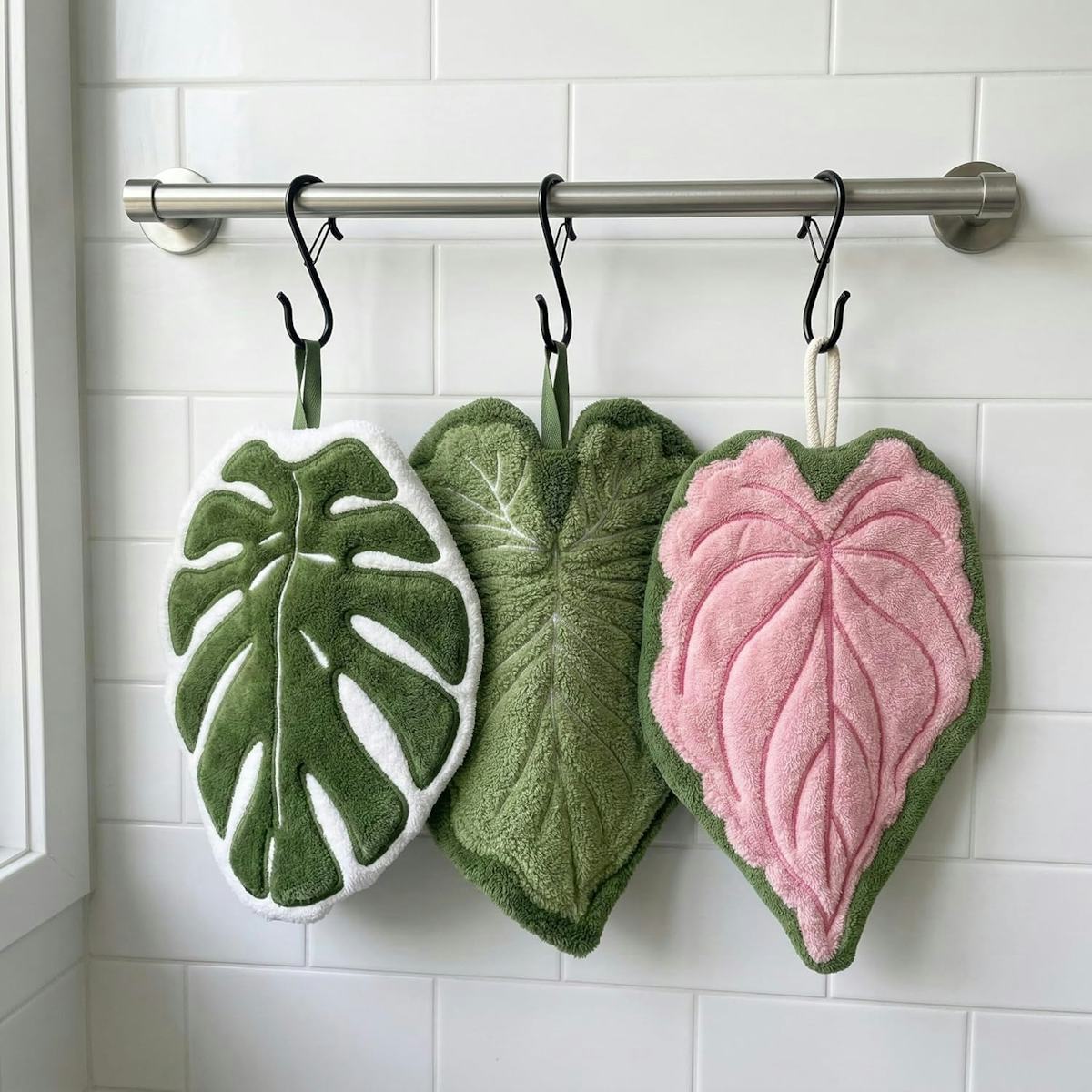 Three plush tropical leaf hand towels in green, white, and pink hang from black hooks on a stainless bar against white subway tile.