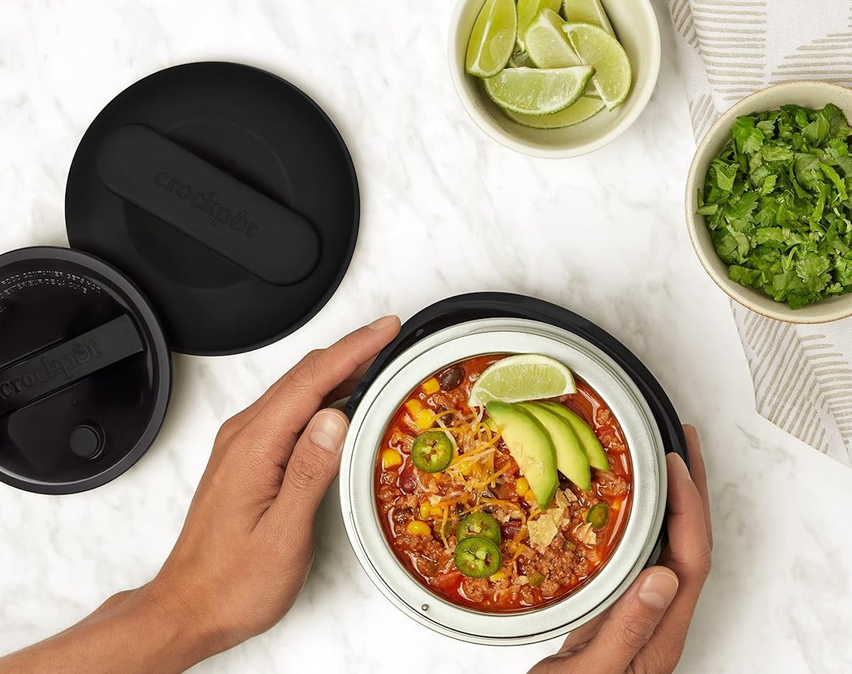 Portable Crock-Pot electric lunch box filled with chili, avocado, lime, cheese, jalapenos, corn, and herbs on a marble counter.
