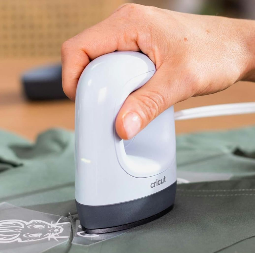 Hand using a Cricut EasyPress Mini portable iron to apply heat-transfer vinyl on green fabric for a DIY craft project.