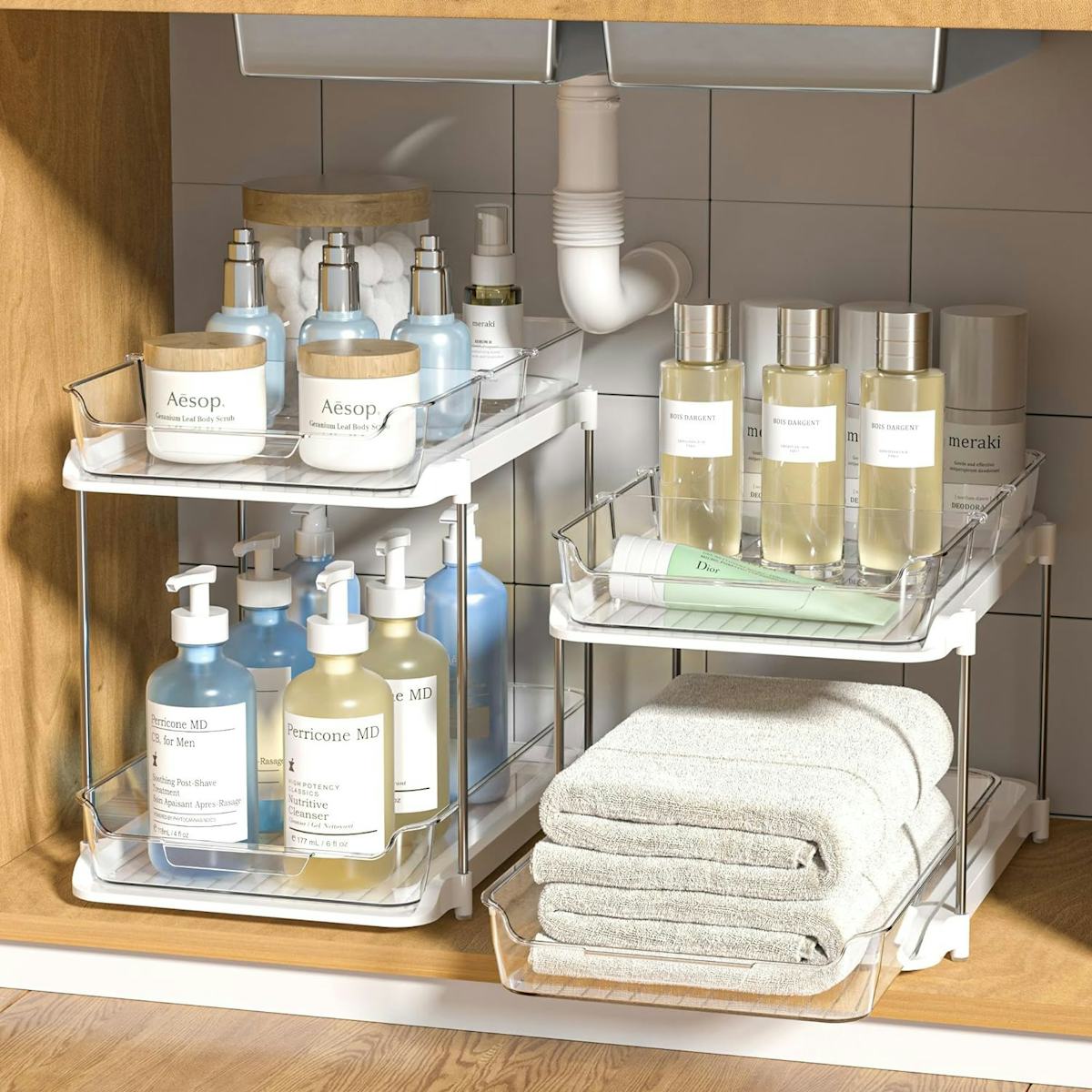 Two clear two-tier under-sink bathroom organizers store skincare bottles, jars, deodorant, and folded towels inside a wood vanity cabinet.