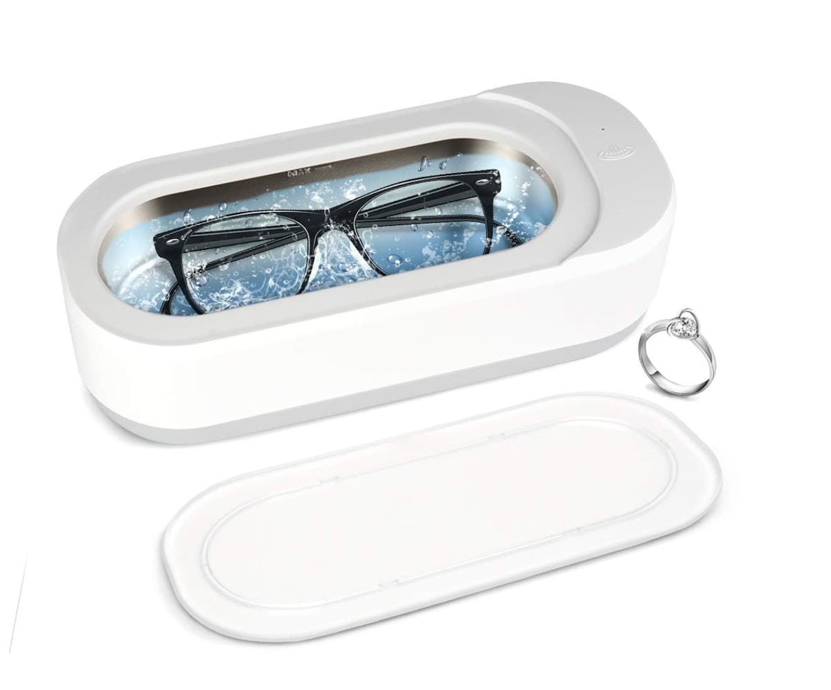 White ultrasonic jewelry cleaner washing eyeglasses in blue water, with a ring beside it and the clear lid placed in front.