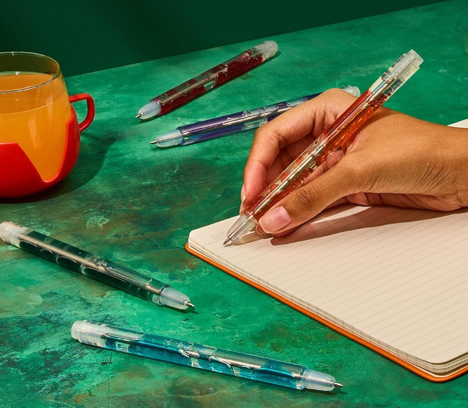 Hand writing in a notebook with a transparent orange lava gel pen, surrounded by colorful gel pens and an orange mug on a green tabletop.