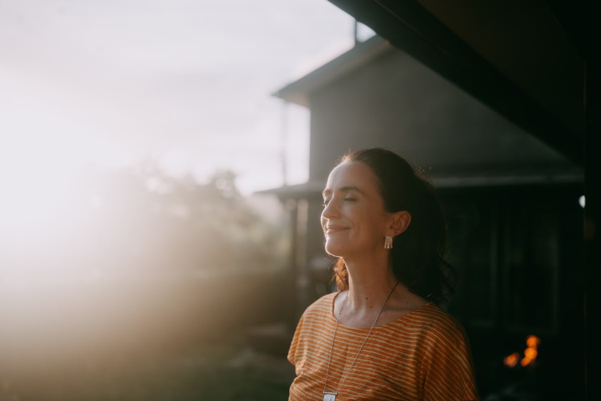Woman with eyes closed smiling in warm sunlight outdoors, wearing a striped orange top and earrings in a calm lifestyle portrait.