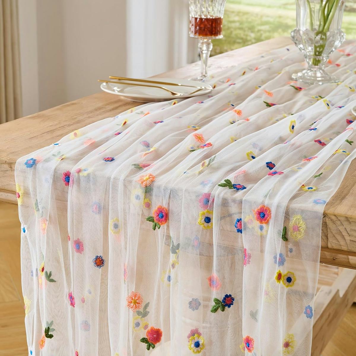 Sheer white embroidered floral table runner draped over a wooden dining table with colorful flowers, plates, gold flatware, glassware, and a vase.