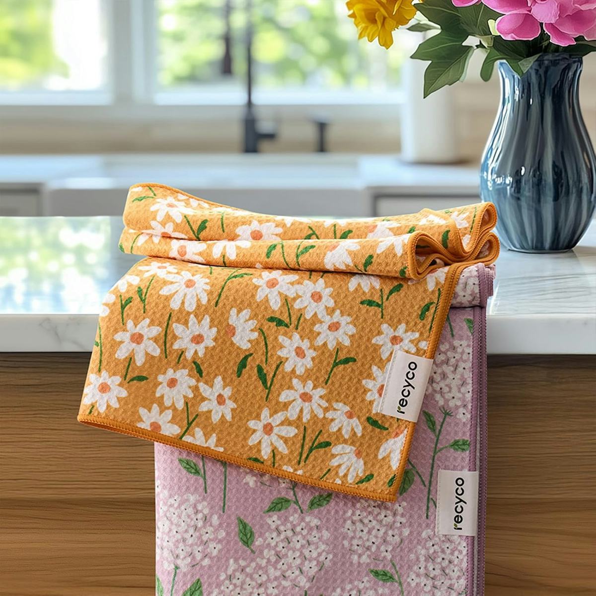 Two floral waffle kitchen towels, one orange with daisies and one pink with tiny flowers, stacked on a counter beside a blue vase.