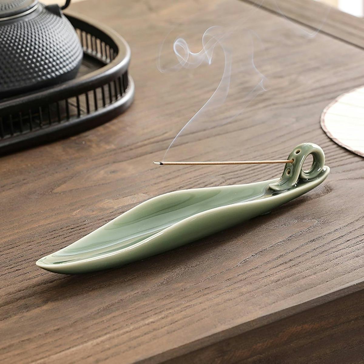 Green ceramic leaf-shaped incense holder with a burning incense stick and wisps of smoke on a wooden tabletop.