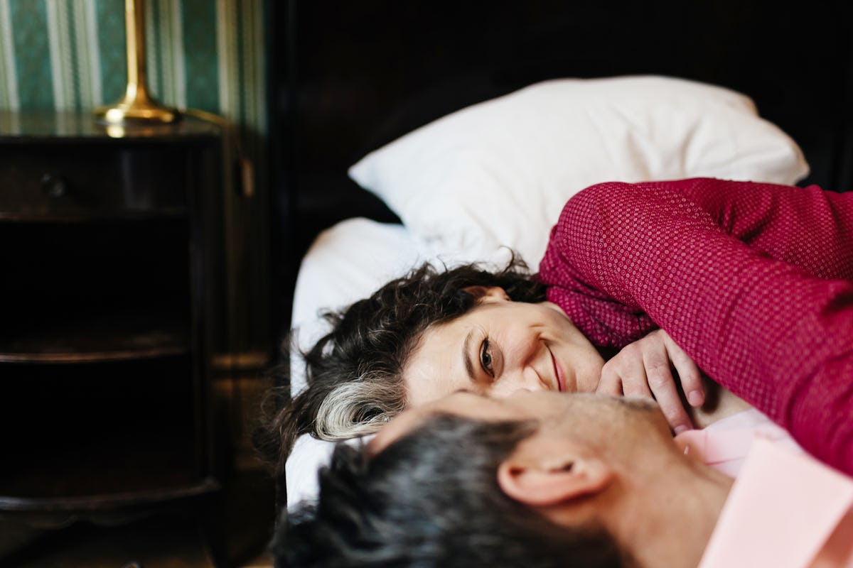 Mature couple lying close together in bed, with a smiling woman looking at her partner under a red blanket and white pillows in a cozy bedroom.