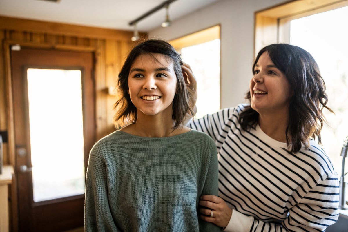 Mother gently touches her smiling teenage daughter's hair in a sunlit home, showing a warm parent-child moment during adolescence.