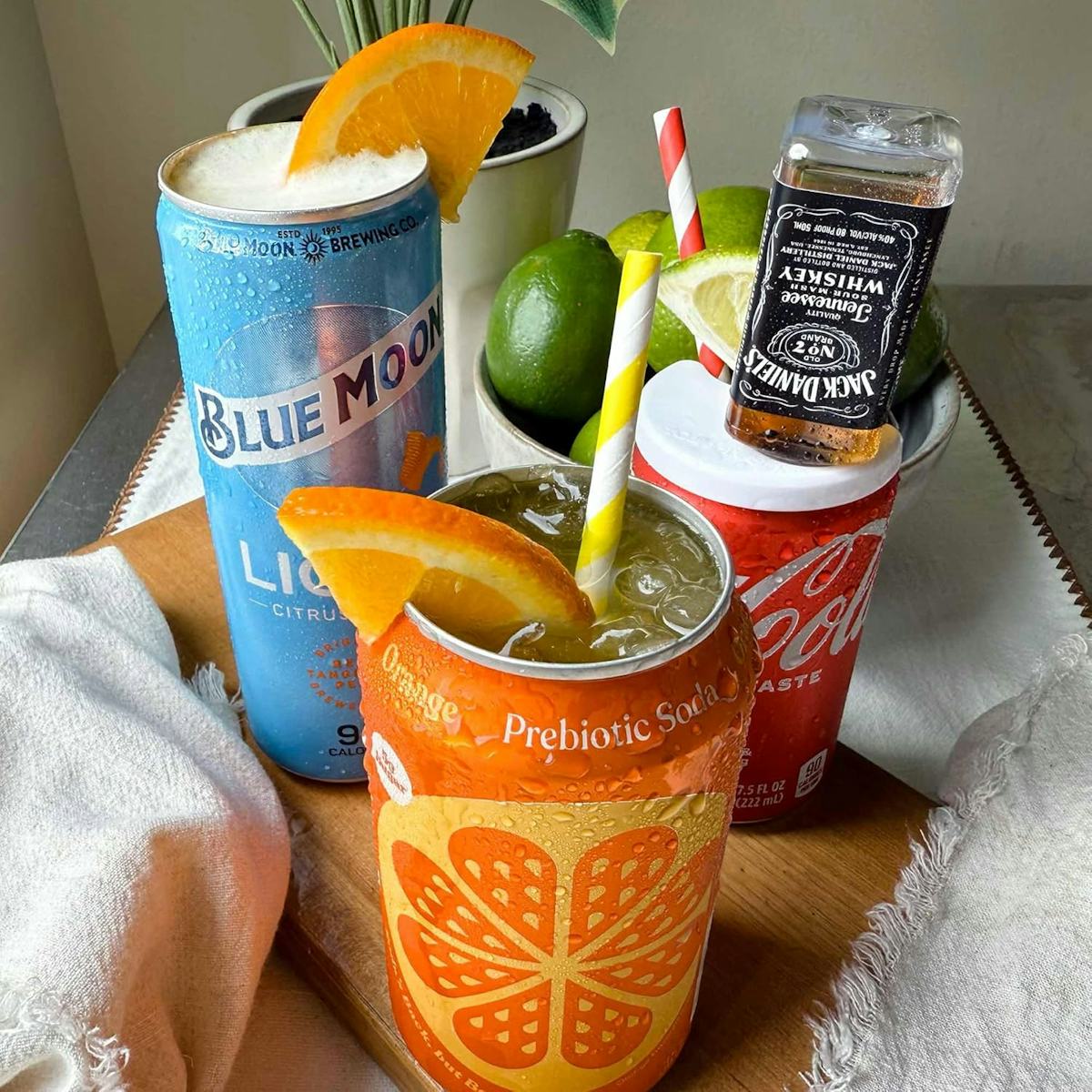 Colorful canned drinks on a wooden tray, including Blue Moon beer, Olipop soda, Coke, citrus slices, limes, straws, and a mini whiskey bottle.