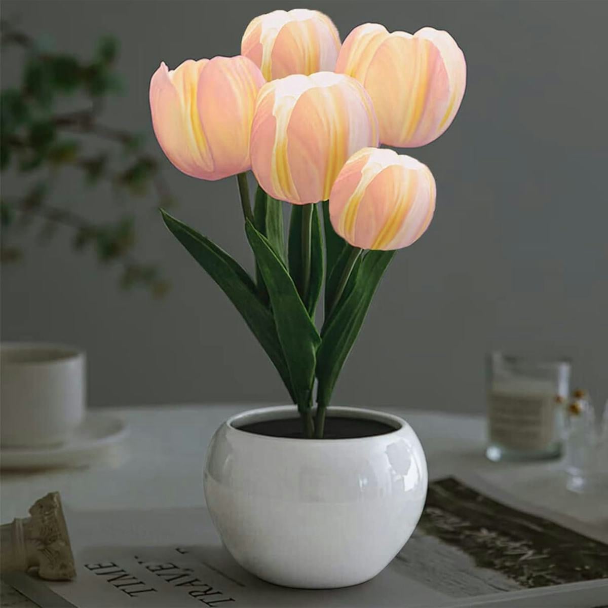 Soft pink LED tulip table lamp in a glossy white pot, displayed on a tabletop with blurred home decor in the background.
