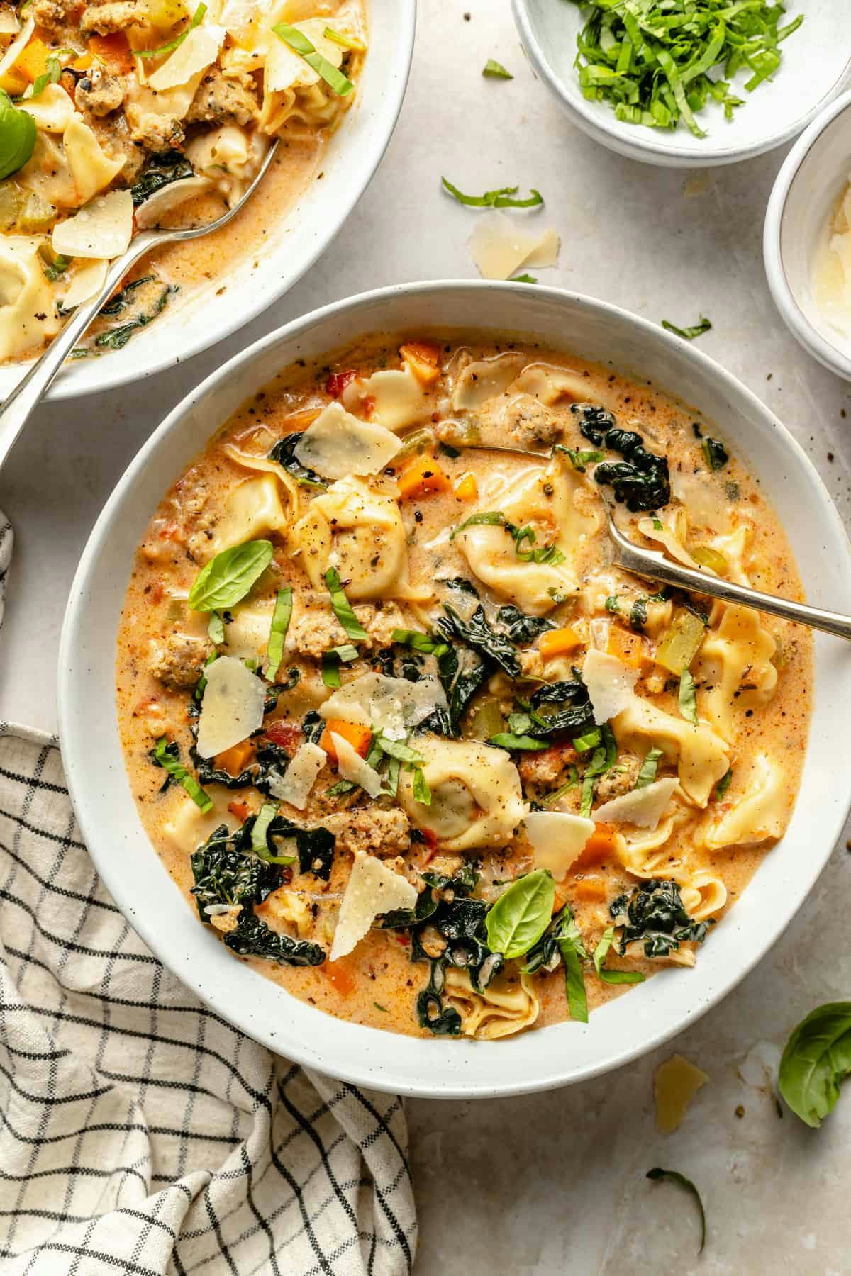 Creamy sausage tortellini soup with kale, carrots, basil, and Parmesan in a white bowl on a marble surface.