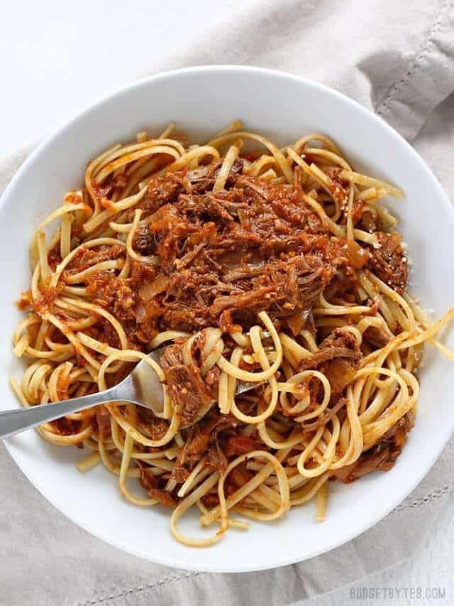 White bowl of spaghetti topped with slow-cooker beef ragu and rich tomato sauce, served with a fork on a light cloth surface.