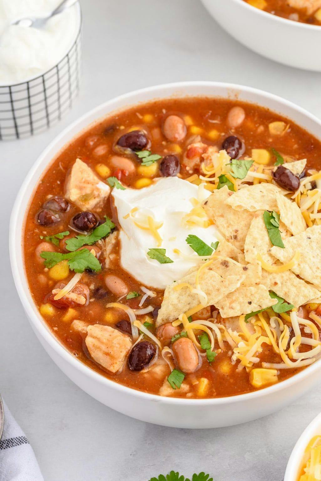 Bowl of tomato-based chicken taco soup with beans, corn, tortilla chips, shredded cheese, sour cream, and cilantro on a light table.