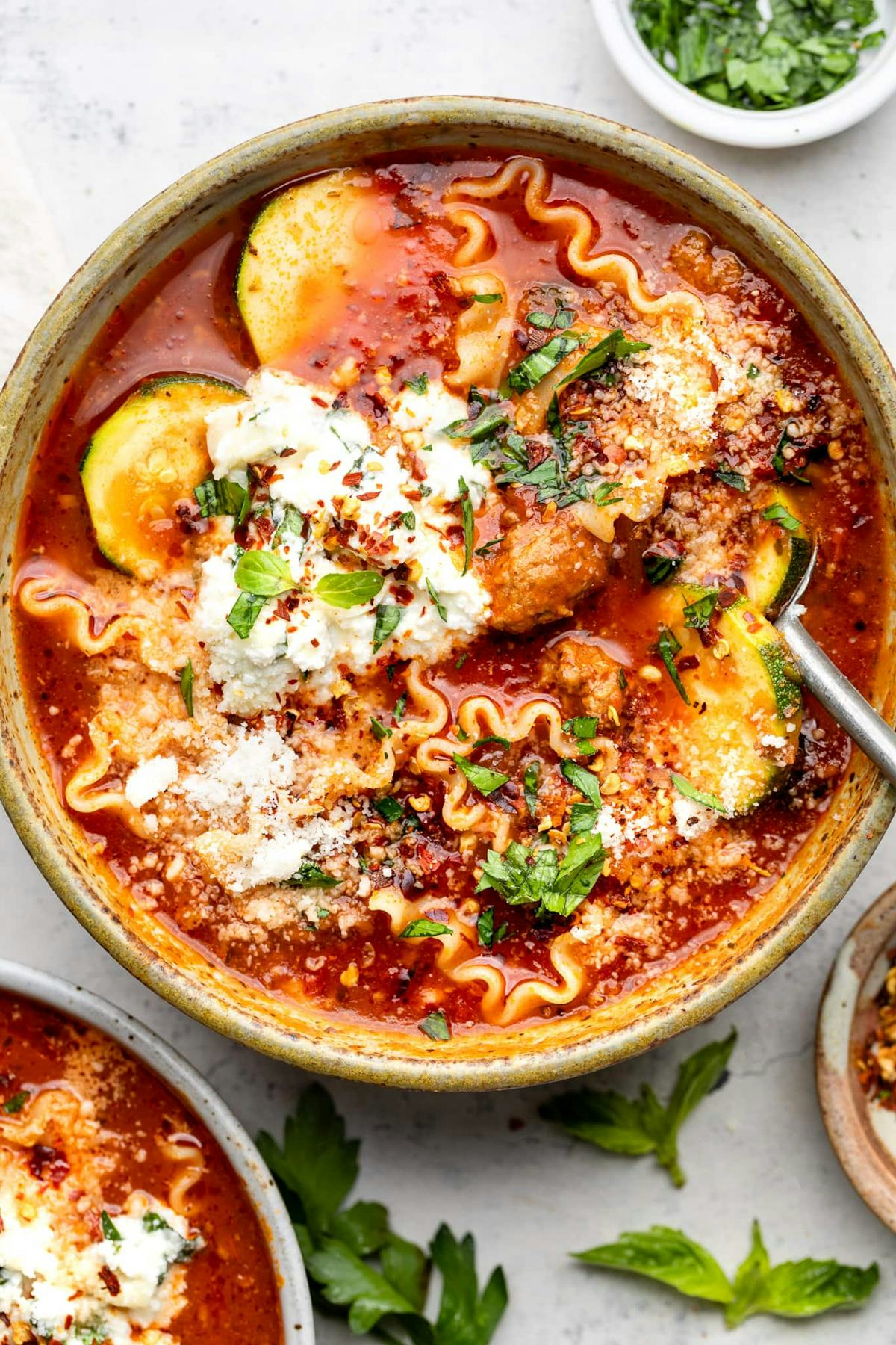 Bowl of lasagna soup with tomato broth, wavy noodles, zucchini, meatballs, ricotta, Parmesan, herbs, and red pepper flakes.