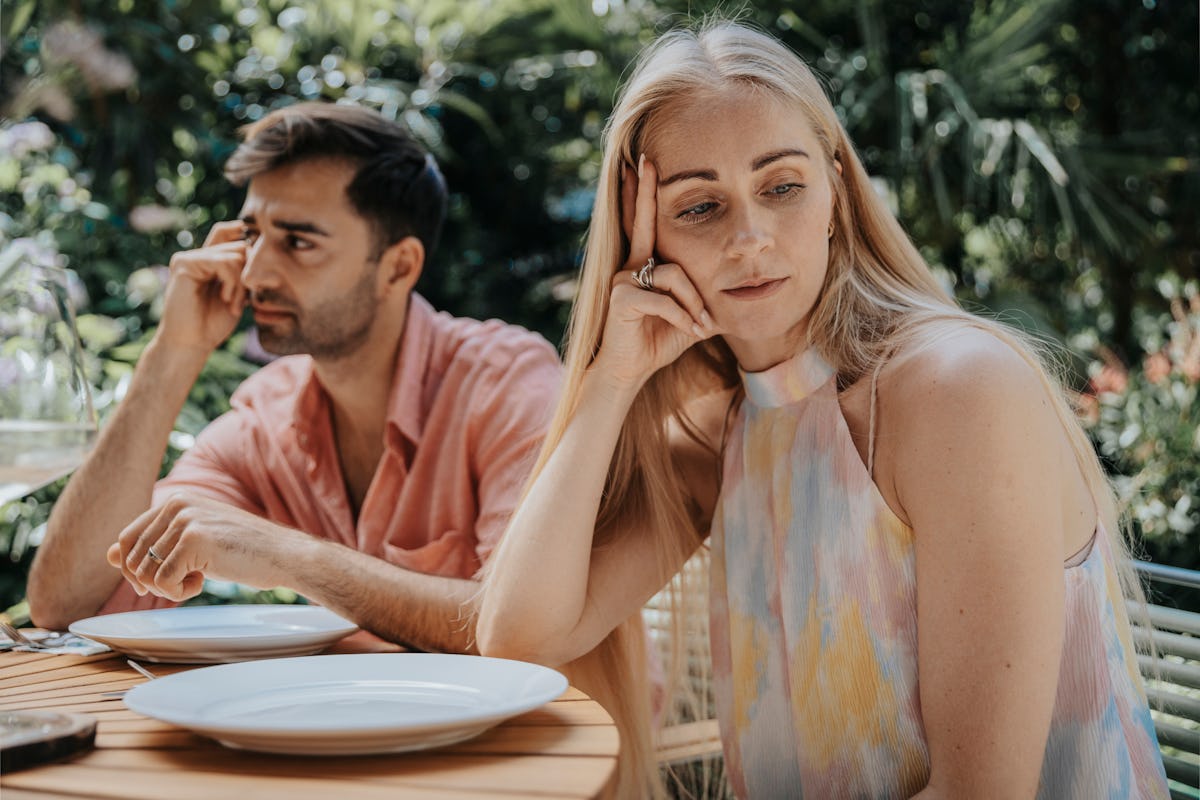 A distant couple sits at an outdoor restaurant table, looking away from each other with tense expressions, suggesting emotional distance in marriage.