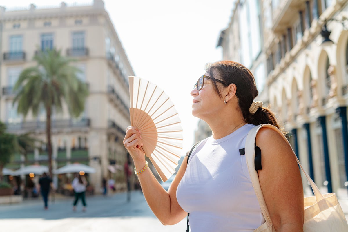 Woman in sunglasses uses a folding fan on a hot sunny city street, illustrating summer heat sensitivity and overheating risk.