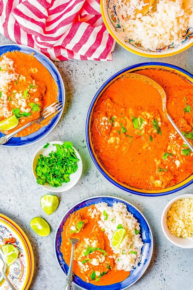 Overhead view of creamy chipotle chicken in tomato sauce served with white rice, cilantro, lime wedges, and queso fresco in blue bowls.