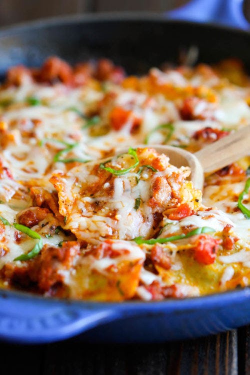 Cheesy ravioli and Italian sausage skillet in tomato sauce, served in a blue pan with a wooden spoon and green herb garnish.
