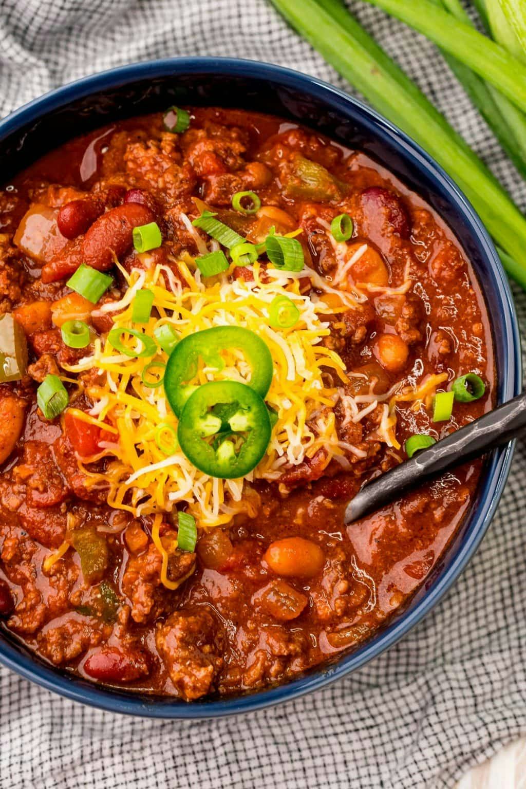Bowl of hearty chili with beans, ground meat, shredded cheese, green onions, and jalapeno slices beside celery sticks.