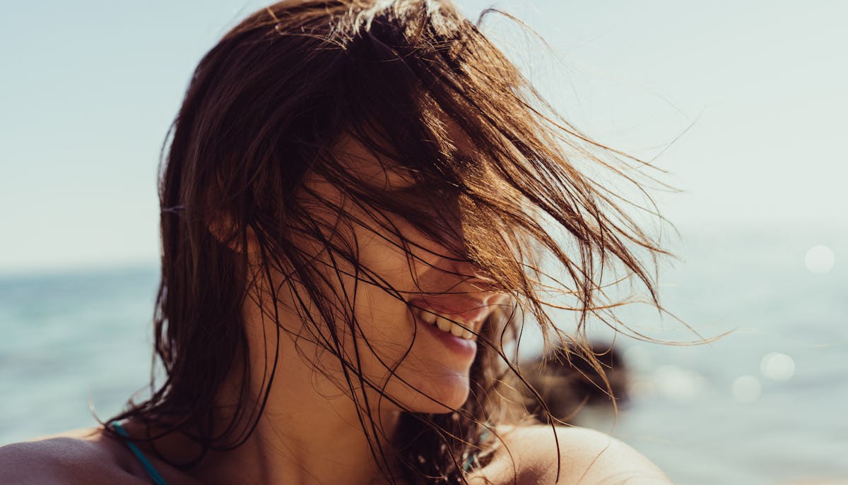Close-up of a smiling woman at the beach with wet windblown hair, bright sunlight, and the ocean softly blurred behind her.