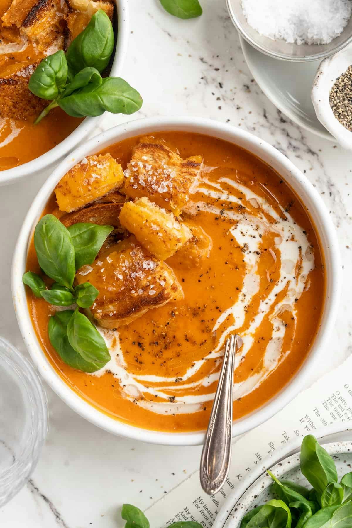 Creamy tomato soup in a white bowl with cheesy croutons, basil leaves, cream swirl, salt, and pepper on a marble surface