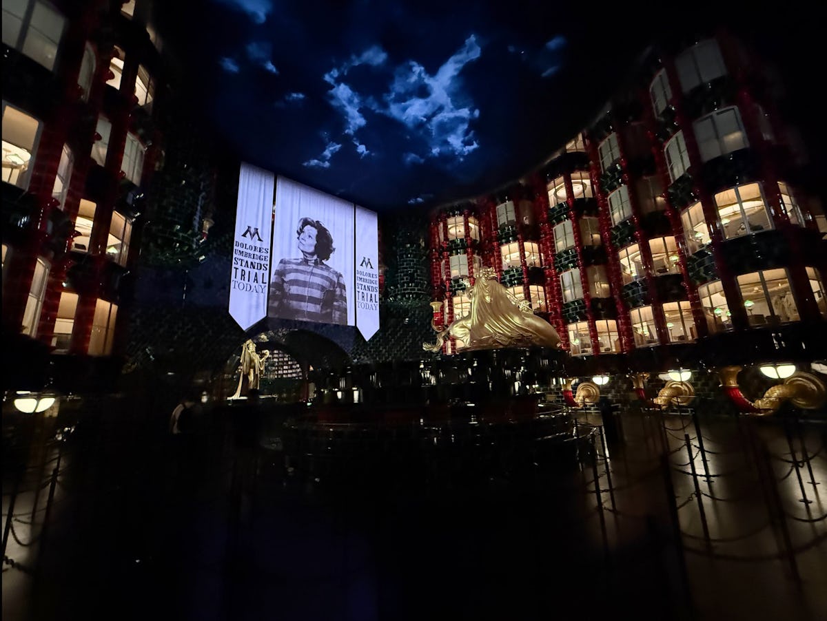 Dimly lit Ministry of Magic atrium at Universal Epic Universe with Dolores Umbridge trial banners, glowing windows, gold statue and stormy ceiling.