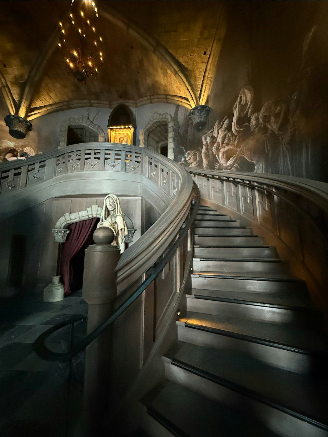 Dimly lit Gothic staircase inside Universal Epic Universe's Dark Universe with stone arches, chandelier, murals, and a fortune teller statue.