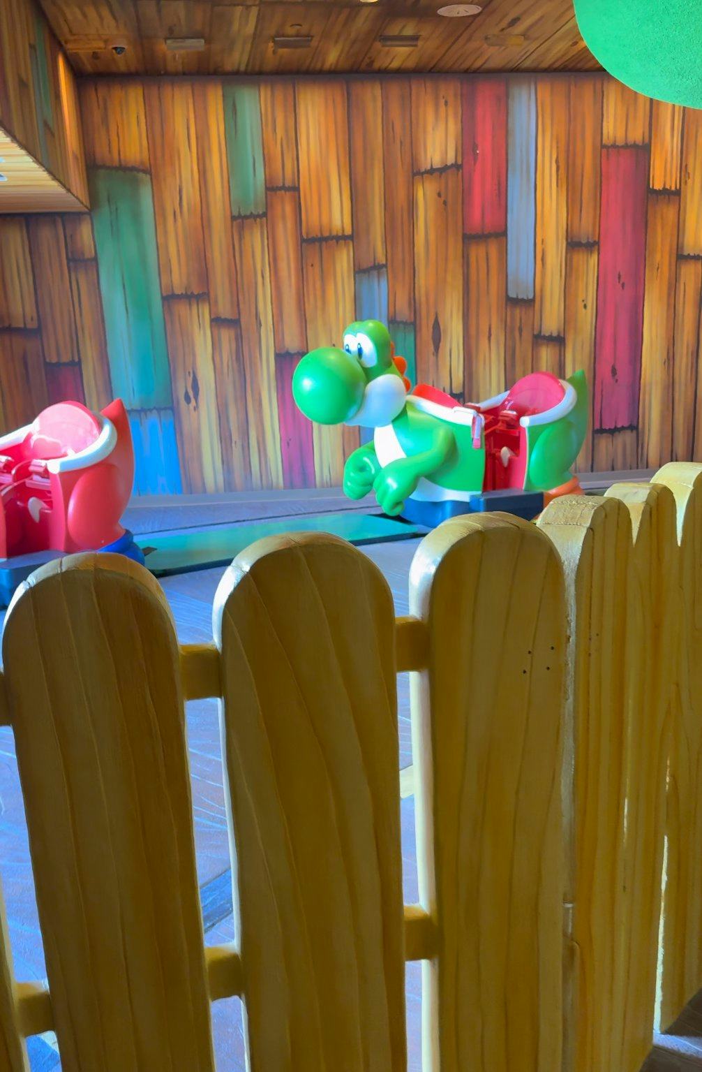 Green Yoshi ride vehicle on Yoshi's Adventure at Super Nintendo World, seen behind a yellow fence and colorful wood-paneled theme park set.