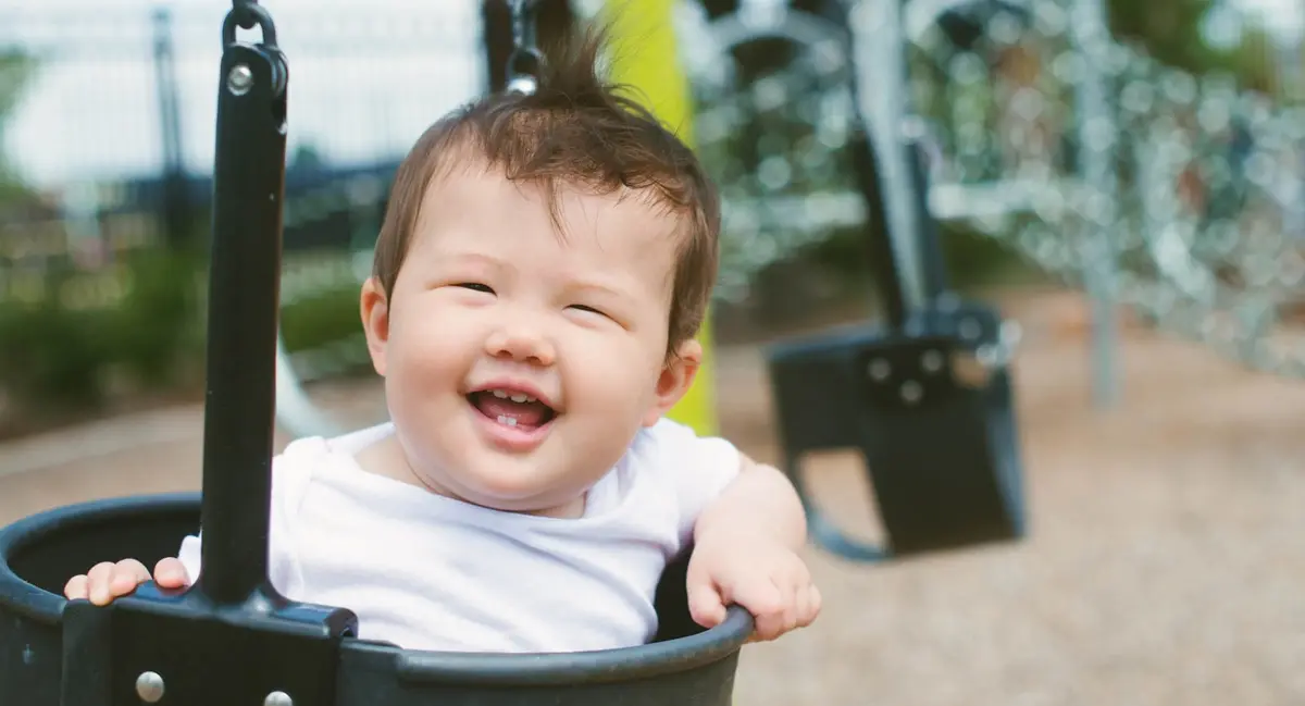 Smiling baby boy in a playground bucket swing, wearing a white shirt with blurred swings and park equipment in the sunny background.