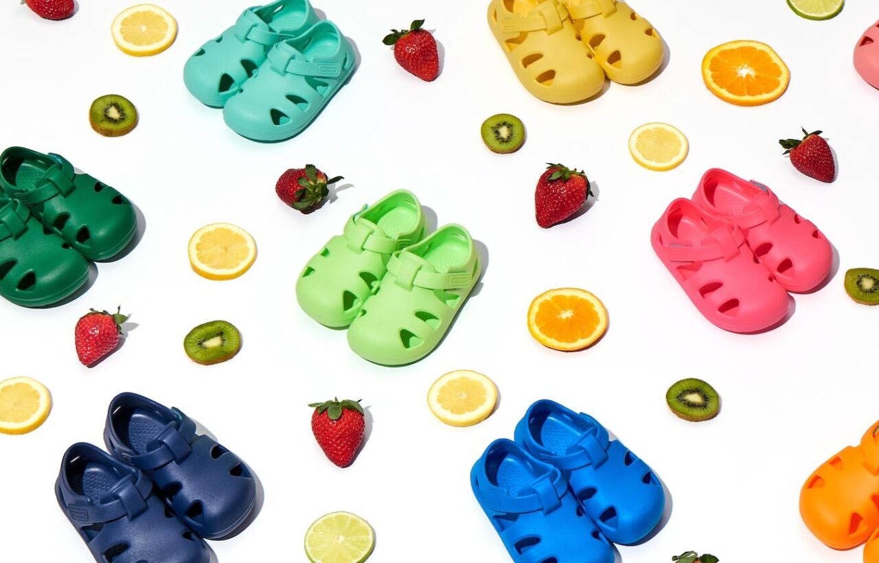 Colorful toddler clogs arranged with citrus slices, strawberries, and kiwi on a bright white background for a playful shoe display.