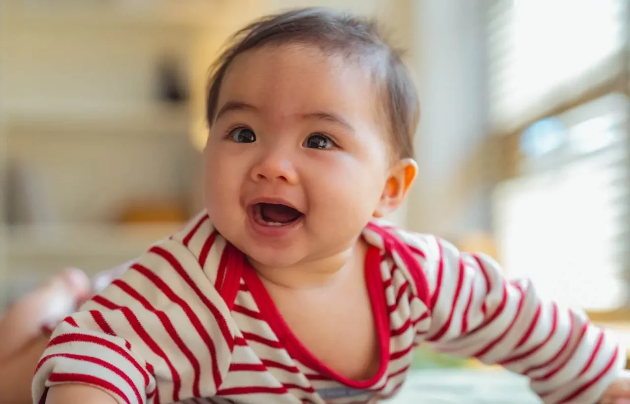 Smiling baby in a red and white striped outfit lying on a bed in soft natural window light.